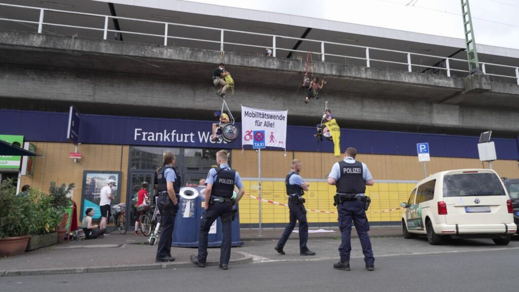 Vor dem Eingang zum Frankfurt West Bahnhof hängen vier Menschen auf Kletterseile, zwei davon im Rollstuhl, mit einem großen Banner "Mobilitätswende für Alle!". Vier Polizist*innen stehen etwas ratlos davor.