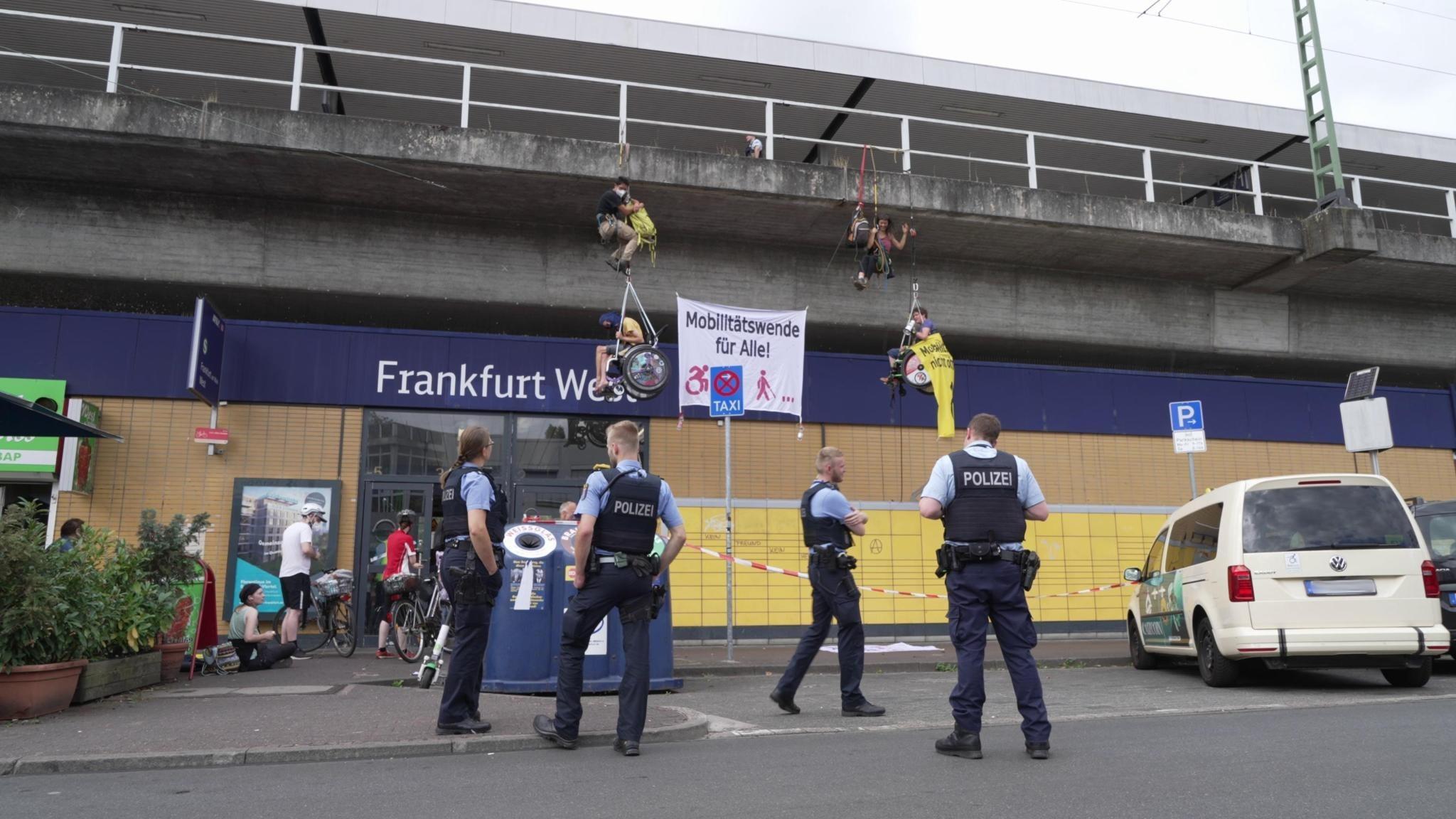 Four people hang from climbing ropes in front of the entrance to Frankfurt West train station. Two of them are in wheelchairs, and between them is a big banner saying "Mobility transition for everyone!" Four police officers are standing on the ground below them.